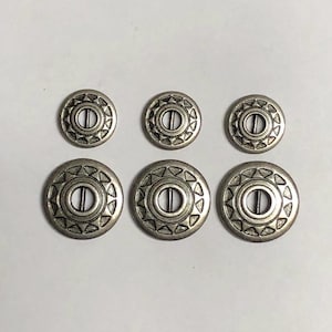 Könnte beinhalten: Ein Set aus sechs runden Metallknöpfen, angeordnet in zwei Reihen zu je drei Stück. Jeder Knopf hat eine silberfarbene Oberfläche mit einem horizontalen Mittelsteg und einem dekorativen Rand aus kleinen, nach innen zeigenden Dreiecken.