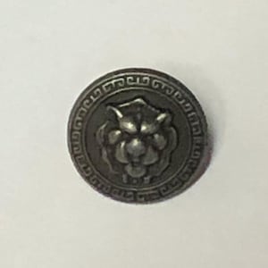 May include: A round, metallic button featuring a raised, embossed lion head at the center. The button has a dark, antiqued finish and is surrounded by a decorative, geometric border pattern that encircles the central lion motif.