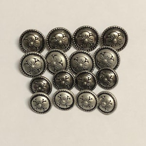 May include: A collection of fifteen round, silver-toned metal buttons arranged in a grid. Each button features a raised, embossed five-petal floral design with a beaded border around the edge, creating a consistent metallic texture across all pieces.