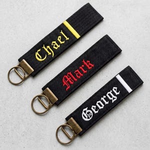 May include: Three black fabric keychains with bronze-toned metal rings. Each keychain features embroidered text in a gothic font: the first reads Chael in yellow, the second reads Mark in red, and the third reads George in white, each with a matching stripe.