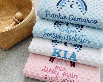 May include: A stack of four personalized baby blankets in shades of blue, white, and pink. Each blanket features a rainbow design with embroidered names and dates. The blankets are soft and textured, perfect for a baby.