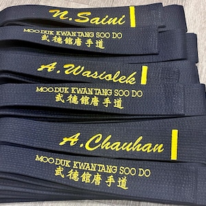 May include: Three black martial arts belts stacked vertically. Each belt features yellow embroidered text reading "MOO DUK KWAN TANG SOO DO" in English and Korean characters, along with a personalised name and a vertical yellow stripe on the right side.