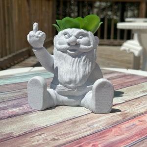 May include: A white ceramic gnome planter with a green plant growing out of its head. The gnome is giving the middle finger.