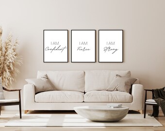 Op de afbeelding: Drie zwarte ingelijste prints met witte achtergronden en zwarte tekst. De prints lezen "I AM Confident", "I AM Fierce" en "I AM Strong". De prints hangen aan een lichtbeige muur boven een witte bank.
