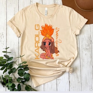 K&ouml;nnte beinhalten: Beiges T-Shirt mit einer Cartoon-Illustration einer Frau mit Federkopfschmuck. Das Wort "KAROLE" ist vertikal auf der linken Seite des Shirts in orangefarbenen Buchstaben aufgedruckt. Die Illustration enth&auml;lt ein Herz-Tattoo und den Namen "Aria" in Kursivschrift.