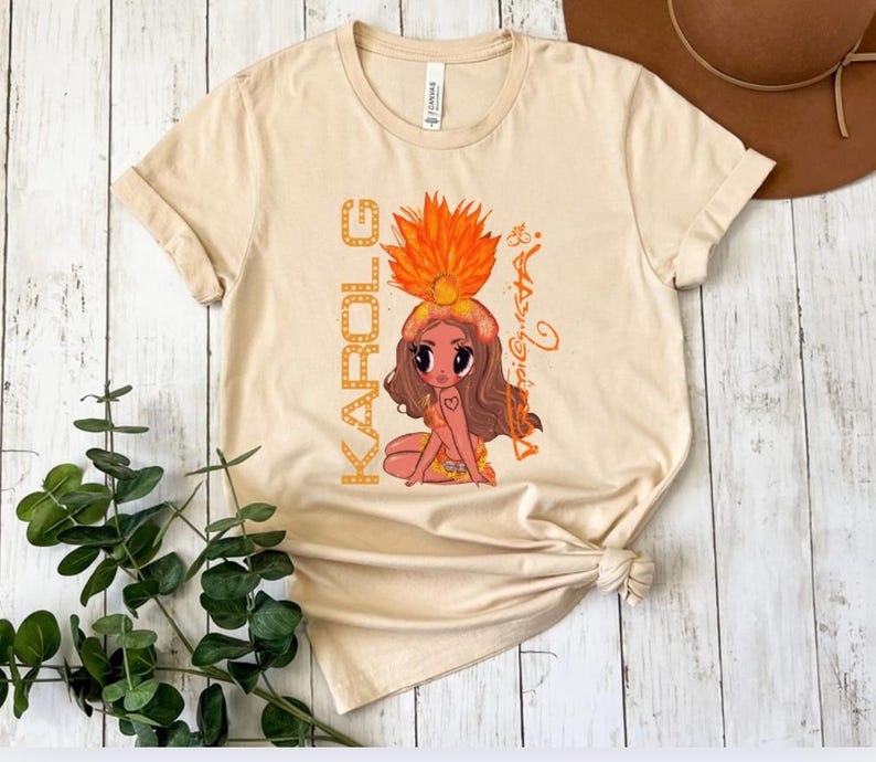 K&ouml;nnte beinhalten: Beiges T-Shirt mit einer Cartoon-Illustration einer Frau mit Federkopfschmuck. Das Wort "KAROLE" ist vertikal auf der linken Seite des Shirts in orangefarbenen Buchstaben aufgedruckt. Die Illustration enth&auml;lt ein Herz-Tattoo und den Namen "Aria" in Kursivschrift.