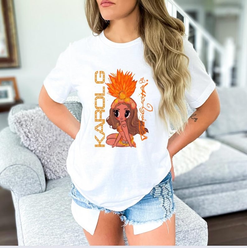 K&ouml;nnte beinhalten: Wei&szlig;es T-Shirt mit einer Cartoon-Illustration eines M&auml;dchens mit Federkopfschmuck und dem Namen "KAROL G" in goldfarbenen Buchstaben. Die Illustration ist im Cartoon-Stil mit einem Herz auf der Brust des M&auml;dchens. Das Shirt wird mit Jeansshorts getragen.