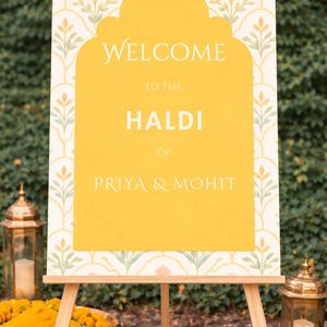 May include: A wooden easel displays a welcome sign for a Haldi ceremony. The sign features a yellow arch shape with the words "WELCOME TO THE HALDI OF PRIYA & MOHIT" in a cream color. The background has a floral pattern.