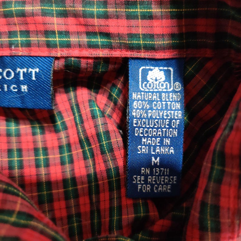 K&ouml;nnte beinhalten: Nahaufnahme eines rot, gr&uuml;n und gelb karierten Hemdes. Das Hemd hat ein blaues Etikett mit dem Text "cotton" und "NATURAL BLEND 60% COTTON 40% POLYESTER EXCLUSIVE OF DECORATION MADE IN SRI LANKA M RN 13711 SEE REVERSE FOR CARE."