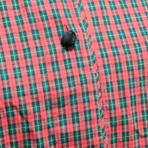 K&ouml;nnte beinhalten: Nahaufnahme eines rot-gr&uuml;nen Stoffes mit einem schwarzen Pompon-Knopf. Der Stoff hat ein klassisches Karomuster mit gr&uuml;nen und goldenen Linien. Der Knopf ist mit einem kleinen roten Faden am Stoff befestigt.