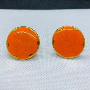 Puede incluir: Un par de pendientes redondos de color naranja y dorado. Los pendientes están hechos de metal y tienen un acabado liso y brillante.