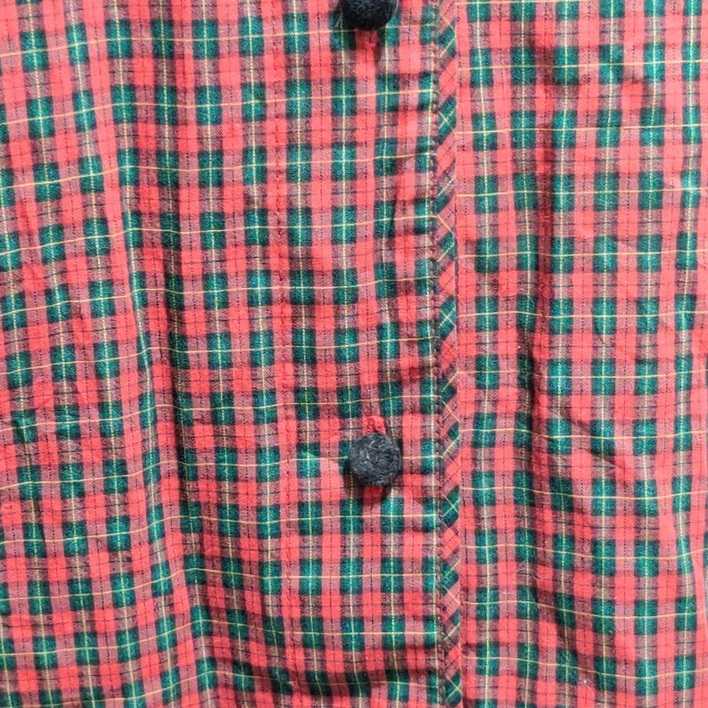 K&ouml;nnte beinhalten: Nahaufnahme eines rot-gr&uuml;nen karierten Stoffes mit Knopfleiste. Der Stoff hat ein klassisches Karomuster mit dunkelgr&uuml;nen und hellbraunen Linien. Die Kn&ouml;pfe sind dunkelgrau.