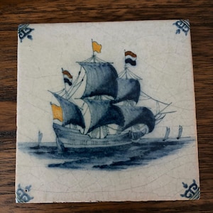 May include: Square ceramic tile featuring a detailed illustration of a sailing ship on a blue sea. The ship has blue sails and flags in orange, white, and black. The tile has a white background with blue corner designs.