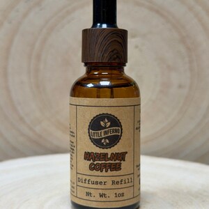 May include: A brown glass bottle with a wooden dropper and a label that reads "Hazelnut Coffee Diffuser Refill." The bottle is 28g and has the "Little Inferno" logo. The bottle is on a wooden surface.
