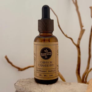 May include: An amber glass bottle with a black dropper cap and a wood-grain collar. The label is tan paper with black text reading Little Inferno Crimson Cranberry Diffuser Refill Nt. Wt. 1oz, featuring a circular logo with leaf icons.