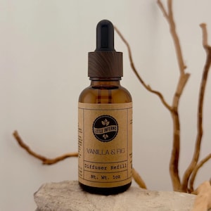 May include: An amber glass bottle with a black dropper cap and a wood-grain textured collar. The label is tan paper with black text reading Little Inferno, Vanilla & Fig, Diffuser Refill, and Nt. Wt. 28g, featuring a circular logo with leaf icons.