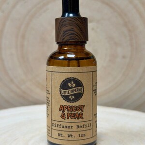 May include: An amber glass dropper bottle with a wood-grain patterned cap. The tan label features the text Little Inferno, Apricot & Pear, Diffuser Refill, and Nt. Wt. 28g. The bottle contains a liquid and has a black rubber dropper top.