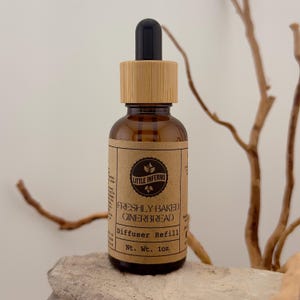 May include: A brown glass bottle with a wooden cap and a black dropper. The label reads "Freshly Baked Gingerbread Diffuser Refill" and "Nt. Wt. 28g". The bottle is set on a light-colored surface with a neutral background.