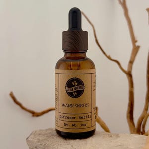 May include: An amber glass bottle with a black dropper cap featuring a wood grain texture. The label is tan paper with black text reading Little Inferno, Warm Winds, Diffuser Refill, and Nt. Wt. 1oz, centred with a circular logo containing leaves.
