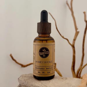 May include: An amber glass bottle with a black dropper cap featuring a wood-grain texture. The tan paper label displays the text Little Inferno, Honey Suckle, Diffuser Refill, and Nt. Wt. 28g, with a circular logo containing three leaf icons.