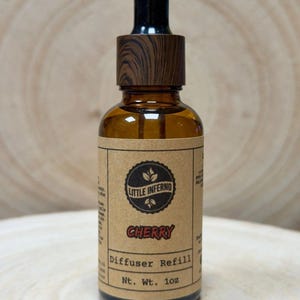 May include: An amber glass dropper bottle with a dark wood-grain patterned cap. The tan paper label features the text Little Inferno, Cherry, Diffuser Refill, and Nt. Wt. 28g. The bottle contains a liquid and is topped with a black glass dropper.