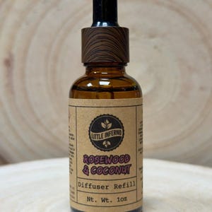 May include: An amber glass dropper bottle with a wood-grain patterned cap. The tan label features the text Little Inferno, Rosewood & Coconut, Diffuser Refill, and Nt. Wt. 28g. The bottle has a black rubber dropper top and contains liquid.