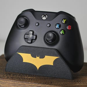 May include: Black Xbox controller on a black stand featuring a gold Batman logo. The controller has coloured buttons and a directional pad. The stand is on a wooden surface.