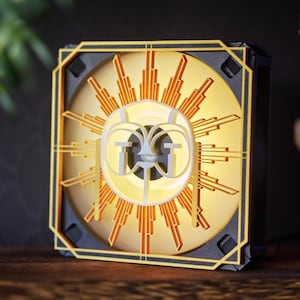 Op de afbeelding: Een vierkante lamp in Art Deco-stijl met een goud en zwart frame. De verlichte binnenkant heeft een sunburst-ontwerp met oranje en witte accenten. Het centrale ontwerp bevat gestileerde letters. De lamp staat op een houten oppervlak.