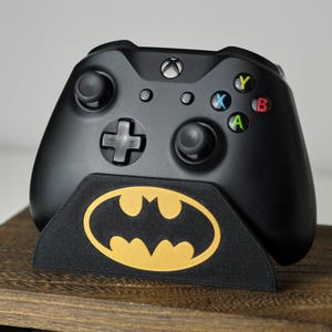 May include: Black Xbox controller on a black stand featuring a gold Batman logo. The controller has coloured buttons and a directional pad. The stand is on a wooden surface.