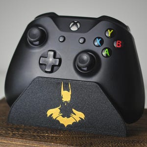 May include: Black Xbox controller with coloured buttons, on a black stand with a gold Batman logo. The controller has two joysticks, a directional pad, and various buttons. The stand is textured and sits on a wooden surface.
