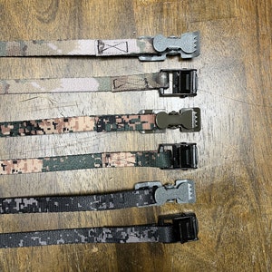 May include: Five different camouflage patterned straps with black and silver buckles. The straps are arranged vertically on a wooden surface.