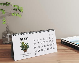 Puede incluir: Calendario de escritorio blanco para mayo con encuadernación en espiral. El calendario presenta una ilustración de una planta y el texto "Let your passion be your guide". El calendario está sobre una superficie de madera.