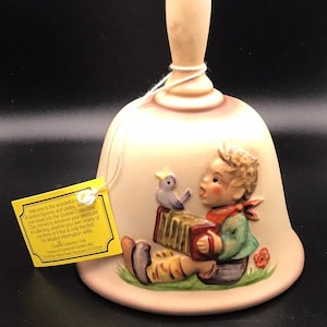 Może przedstawiać: Dzwonek ceramiczny z beżową glazurą i brązowym uchwytem. Dzwonek przedstawia płaskorzeźbę chłopca grającego na akordeonie z niebieskim ptakiem siedzącym na górze. Dzwonek ma przymocowaną żółtą etykietę z tekstem o Goebel Collectors Club.