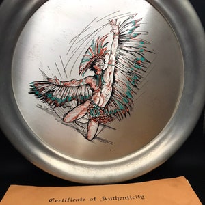 Pode incluir: Um prato oval de prata com uma borda elevada apresenta uma ilustração colorida de um dançarino nativo americano com um cocar de penas. O dançarino é retratado em uma pose dinâmica com os braços estendidos e um cocar de penas que flui. O prato é assinado "Phil Coleman" no canto inferior esquerdo. O prato é acompanhado por um certificado de autenticidade da Master Engravers of America.