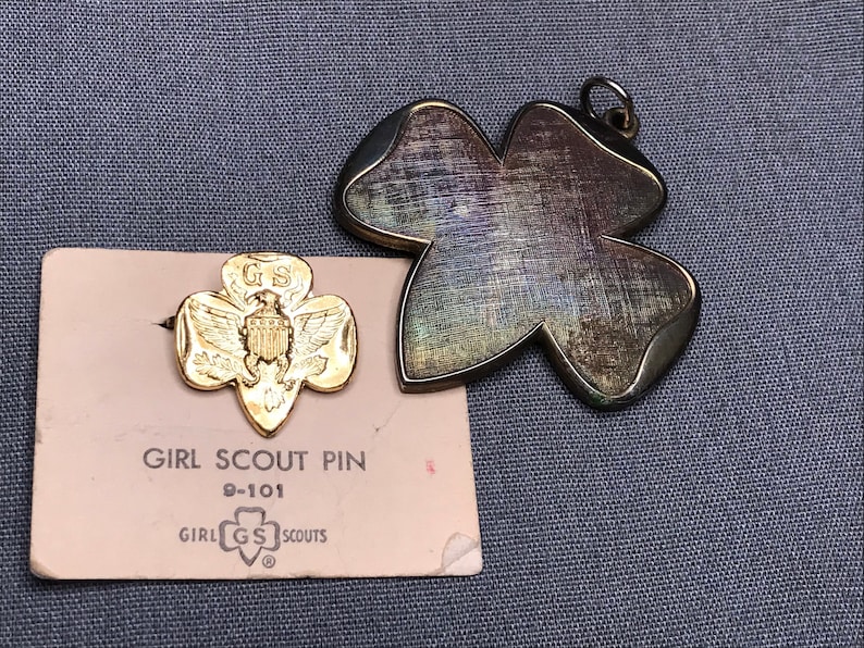 May include: A gold Girl Scout pin with the Girl Scout logo and the words "Girl Scout Pin" and "Girl Scouts" on a pink card. The pin is a gold clover with an eagle and the letters "GS" on it. The card also has the number "D-101" printed on it.