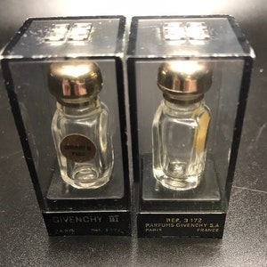 May include: Two vintage Givenchy III perfume bottles in clear glass with gold-coloured lids, presented in black plastic display cases. The cases have clear plastic tops and the Givenchy logo. The bottles have labels that read "Givenchy III Paris".