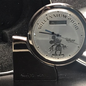 May include: Silver-toned Mickey Mouse clock with a white face and black hands. The clock has a digital display and says "Millennium 2000" and "Disney Quartz".