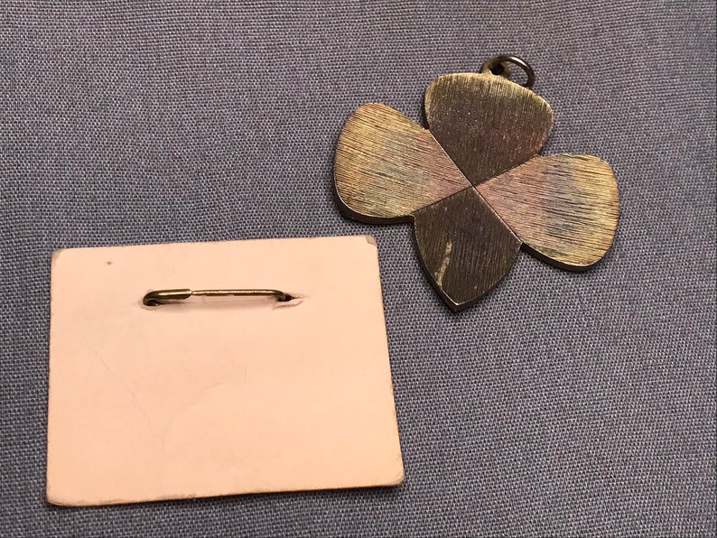 May include: A gold-toned metal pin in the shape of a four-leaf clover. The pin has a safety pin closure and is on a pink backing card.