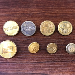 May include: A collection of vintage metal buttons with embossed text. The buttons are in various sizes and colors, including gold and silver. Text includes "Pullman", "Boston Terminal", "Katy", and "Railway Pod Mail Service".