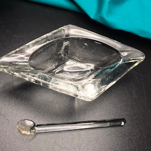 Puede incluir: Un cenicero cuadrado de vidrio transparente con una pequeña cuchara de vidrio. El cenicero está sobre una superficie negra.