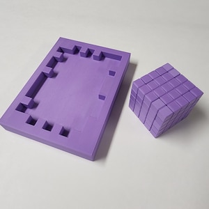 Puede incluir: Dos formas geométricas moradas: un marco rectangular con un diseño pixelado y un cubo compuesto por bloques más pequeños. Los objetos son de un tono morado uniforme y están sobre una superficie blanca. El cubo mide unos 10 cm de ancho.