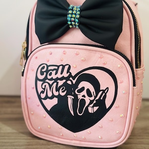 Può includere: Zaino rosa con fiocco nero e strass. La parte anteriore presenta una grafica nera di una faccia di fantasma e le parole "Call Me" all'interno di un cuore. La borsa ha una chiusura con cerniera e piccole perline iridescenti.