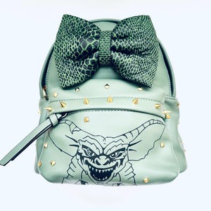 Puede incluir: Una mochila de imitación de cuero verde con tachuelas doradas y una ilustración en blanco y negro de una criatura con cuernos y una boca grande. La mochila tiene un gran lazo verde en la parte superior.