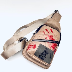 Peut inclure: Un sac à bandoulière en cuir beige avec un motif d'empreinte de main rouge et un patch noir avec un personnage de dessin animé et le texte "Trick or Treat".