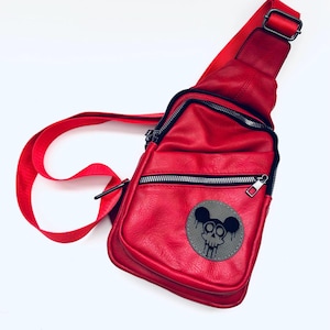 Könnte beinhalten: Eine rote Leder-Umhängetasche mit einem schwarzen Totenkopf- und Mickey-Mouse-Ohren-Design auf der Vorderseite. Die Tasche hat einen Reißverschluss und einen schwarzen verstellbaren Riemen.
