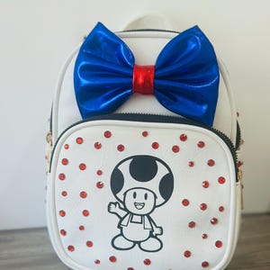 Könnte beinhalten: Weißer Rucksack mit einem Cartoon-Pilz-Charakter-Design. Er hat eine große blaue Schleife mit roter Mitte und ist mit roten Strasssteinen verziert. Die Tasche hat einen Reißverschluss und goldfarbene Hardware.