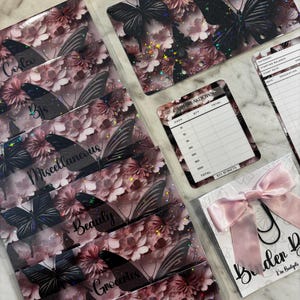 May include: A collection of budget envelopes and accessories. The envelopes feature a floral and butterfly design in shades of pink and black. Categories like "Groceries" and "Beauty" are labeled on the envelopes. A pink bow and paperclip are also included.