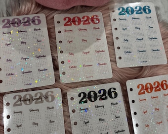 May include: A collection of 2026 planner inserts, each with a calendar layout. The year is displayed in different colours on each insert. The inserts have a holographic glitter effect and are designed with a hole-punched edge for easy insertion.
