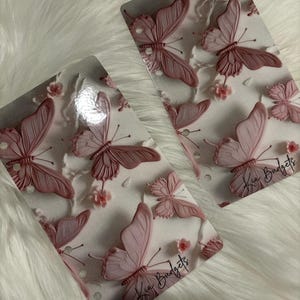 May include: Two clear, rectangular sheets with a pink butterfly and floral design. The butterflies are in various sizes and shades of pink. The text "Kia Budgets" is written in black script on the bottom right corner of each sheet.