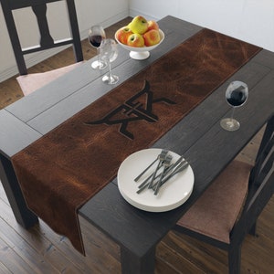 Op de afbeelding: Een bruine leren tafelloper met een zwart logo-ontwerp, op een donkere houten tafel. De tafel is gedekt met witte borden, bestek, wijnglazen en een kom met appels. De loper voegt een rustieke touch toe aan de eethoek.